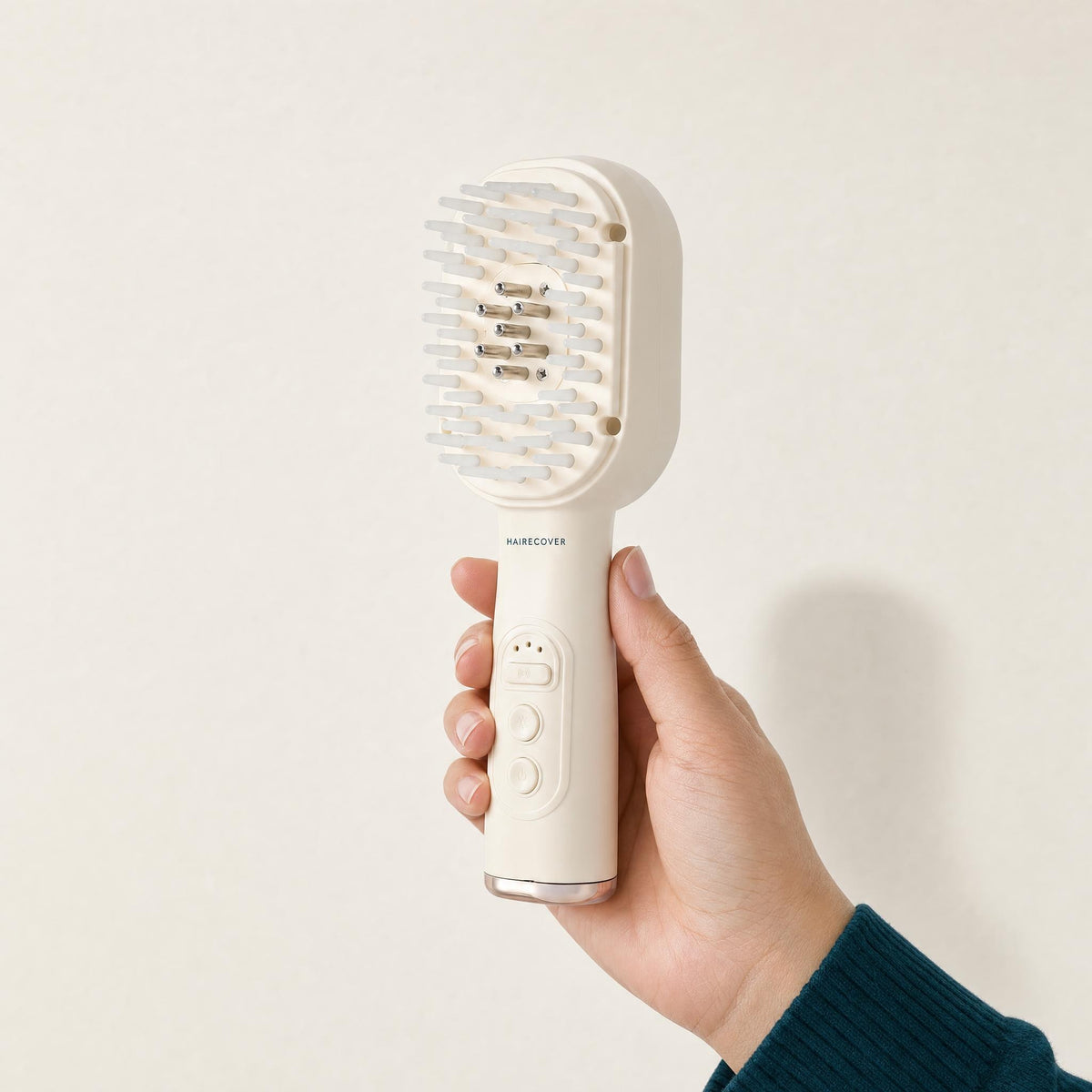 Hand holding the HAIRECOVER cream scalp brush to show its size, on a warm stone background