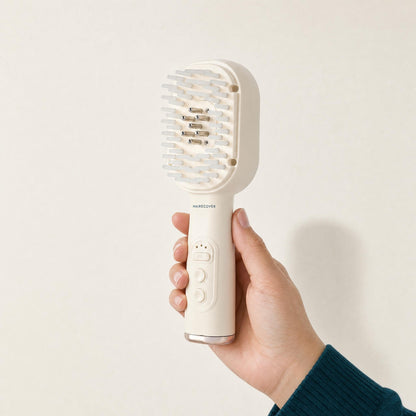 Hand holding the HAIRECOVER cream scalp brush to show its size, on a warm stone background