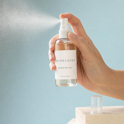 Hand pressing the sprayer of the HAIRECOVER Rosemary Root Tonic bottle, releasing a fine mist in window light